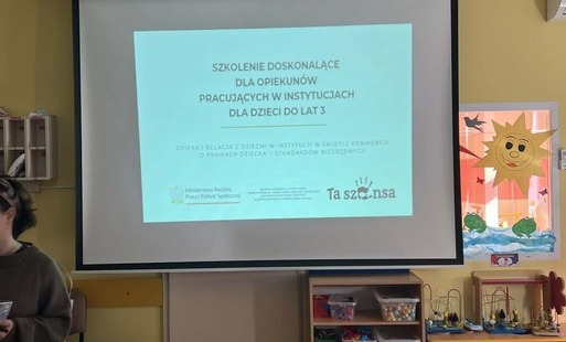 Grafika przedstawia białą tablicę z czarnym napisem "Szkolenie doskonalące dla opiekunów pracujących w instytucjach dla dzieci do lat 3".