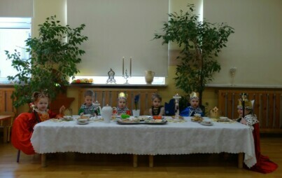 Sala zabaw-na zdjęciu długi stół, na nim biały obrus, zastawa porcelanowa i talerze z jedzeniem. Wokół niego siedzi grupa dzieci ubrana w stroje królewskie.